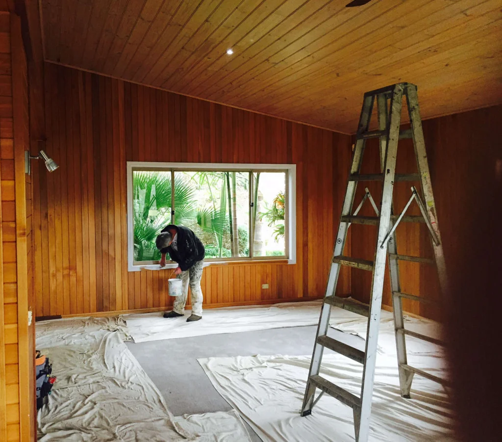 Painter on a ladder in a timber-lined room with floors protected by drop sheets